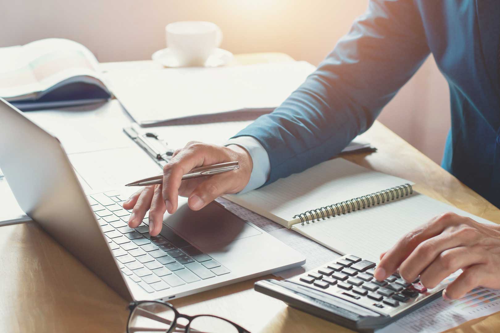 Person schreibt mit Stift und tippt gleichzeitig auf Laptop, daneben Taschenrechner.