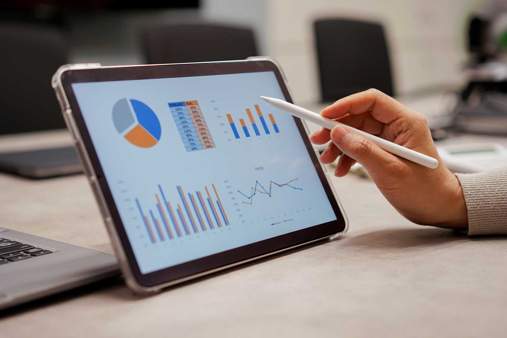 Hand zeigt mit Stift auf Tabletbildschirm mit Diagrammen und Statistiken.