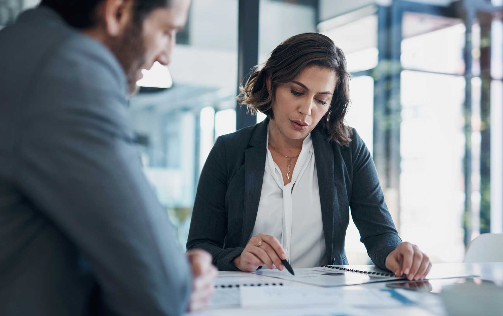 Frau im Business-Outfit zeigt auf Dokumente, Gesprächspartner hört zu.